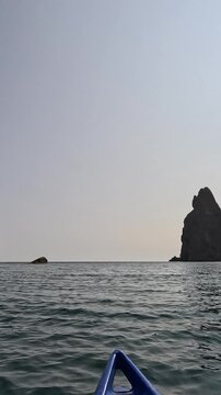 Kayaking past jagged monoliths at sea under bright sunlight. Smooth panning shot reveals impressive rock formations from a small boat perspective.