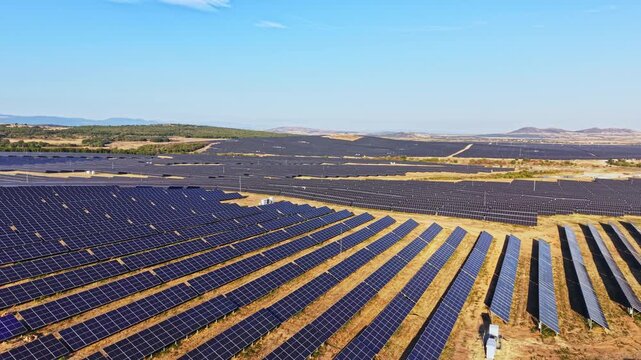 A large solar panel field is located in a rural area. Rows of solar panels are aligned neatly under a clear blue sky. The landscape includes distant hills and open land.