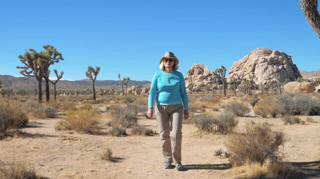 Confident woman wearing hat and sunglasses is walking toward the camera on dirt trail in hot mojave desert, surrounded by iconic joshua trees and large rock formations. Huge boulders and beige rocks.