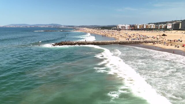 Onshore waves breaking into foamy surf on crowded sandy beach beside rocky jetty and waterfront buildings under bright midday sun