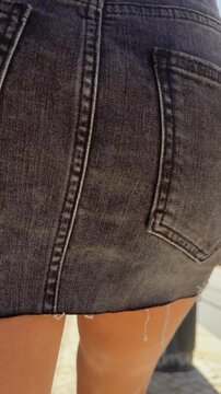 Low-angle street walk of tanned legs in frayed dark denim mini skirt holding smartphone on sunlit urban street