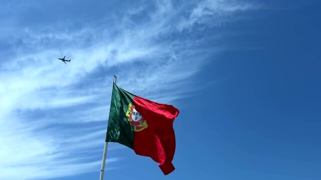 Low-angle view of national flag fluttering in strong wind, red and green fabric with central yellow emblem and partial white shield, waving across bright blue sky with soft clouds