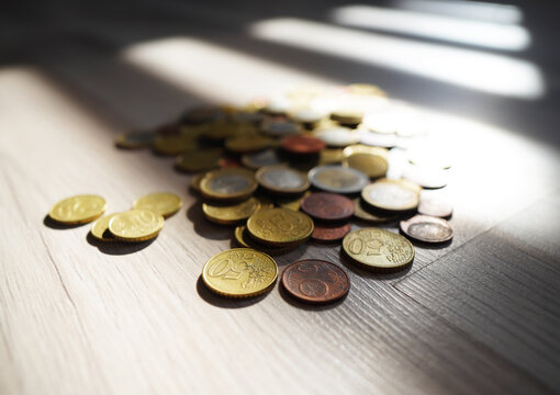 Euro coins scattered on light wood surface with dramatic window light casting sharp shadows and highlights
