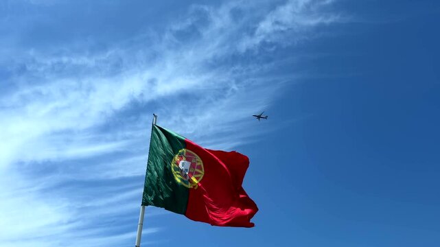 Low-angle view of national flag fluttering in strong wind, red and green fabric with central yellow emblem and partial white shield, waving across bright blue sky with soft clouds
