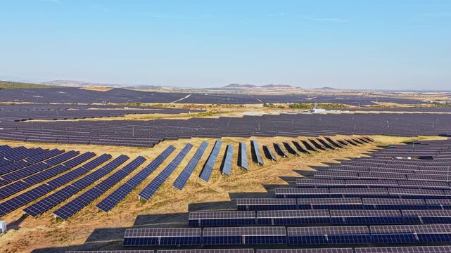 A large solar panel field stretches across the land with rows of panels aligned in straight patterns. The landscape features hills in the distance and clear blue skies.