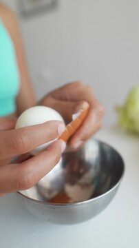 Close-up hands peeling brown eggshell to uncover white hard-boiled egg above stainless steel mixing bowl in bright home kitchen on clean white countertop with steady careful fingers