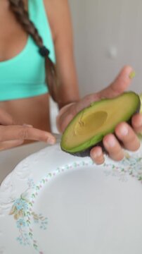 Young woman scooping creamy avocado flesh with spoon from halved fruit over floral ceramic plate, turquoise sports top and braided hair visible in close kitchen view