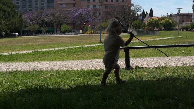Child holding stick at city park edge exploring sunlit lawn from shaded low viewpoint, cobblestone path and metal bench with purple blossoms in calm urban daylight