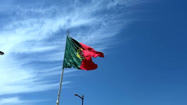 Low-angle view of national flag fluttering in strong wind, red and green fabric with central yellow emblem and partial white shield, waving across bright blue sky with soft clouds