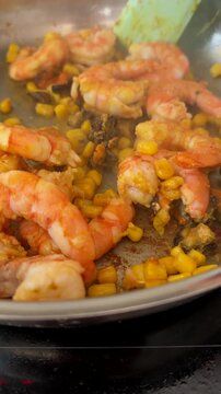 Overhead view of spatula stirring cooked shrimp, corn, and mussels in reflective stainless steel pan with glossy shells and browned sear