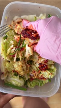 Close-up top-down view of pink gloved hand pressing and tossing spicy napa cabbage salad with red chili paste in clear takeout container on wooden table during meal prep