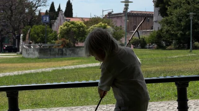 Backlit toddler silhouette manipulating long stick beside shaded pavilion in urban park, standing on grassy lawn with side profile view