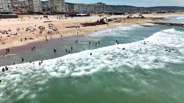 Busy coastal surf zone with whitewater breakers, swimmers and beginner surfers on small surfboards, viewed from elevated oblique angle
