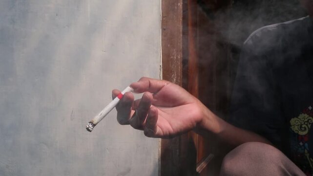 A Javanese man sits casually in front of his house, smoking a kretek clove cigarette during a quiet moment at the doorway.