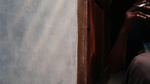 A Javanese man sits casually in front of his house, smoking a kretek clove cigarette during a quiet moment at the doorway.