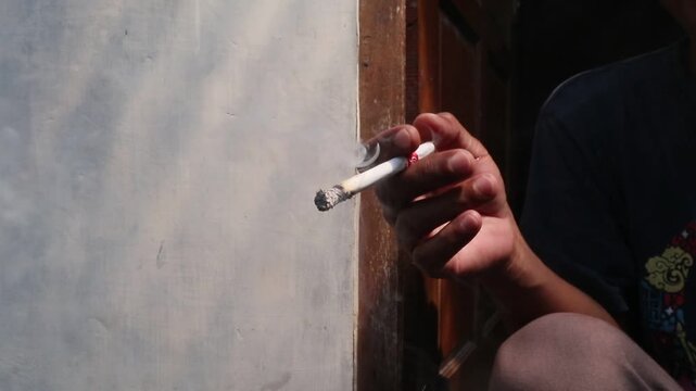A Javanese man sits casually in front of his house, smoking a kretek clove cigarette during a quiet moment at the doorway.