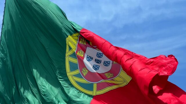 Low-angle view of national flag fluttering in strong wind, red and green fabric with central yellow emblem and partial white shield, waving across bright blue sky with soft clouds