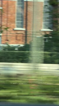 Urban railway roadside pass-by from moving viewpoint with motion-blurred tracks, metal mesh fence, and concrete overpass pillar in bright daylight, vertical framing