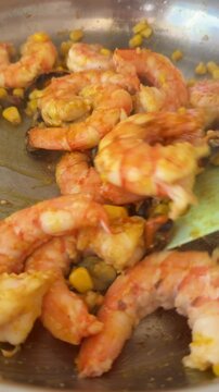 Low angle closeup of hands using silicone spatula to stir and rotate cooked shrimp and corn in reflective stainless steel pan with warm kitchen light and soft steam haze