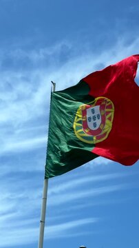 Low-angle view of national flag fluttering in strong wind, red and green fabric with central yellow emblem and partial white shield, waving across bright blue sky with soft clouds