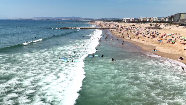 Crowded coastal beach scene with surfers waiting in nearshore waves under bright midday sun, turquoise surf, colorful boards, and swimmers wading near shore by promenade