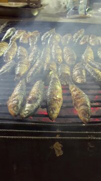 Rows of whole fish on metal grill over glowing charcoal embers, searing to crisp golden skins with rising smoke, glossy sheen, and visible char marks in warm light