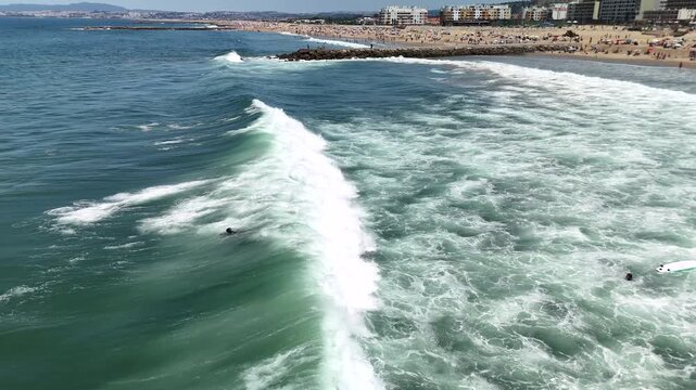 Aerial view of surfers waiting near breaking waves on crowded sandy beach under bright midday sun with umbrellas and low-rise buildings along coastline
