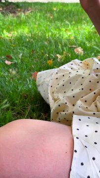 Ground-level close-up of polka-dot skirt and bare thigh as arm with tattoo opens and handles dark denim bag on grassy lawn during picnic leisure in soft daylight