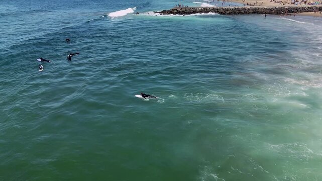 Aerial surf scene of surfers paddling and waiting near rocky jetty over sunlit green blue water