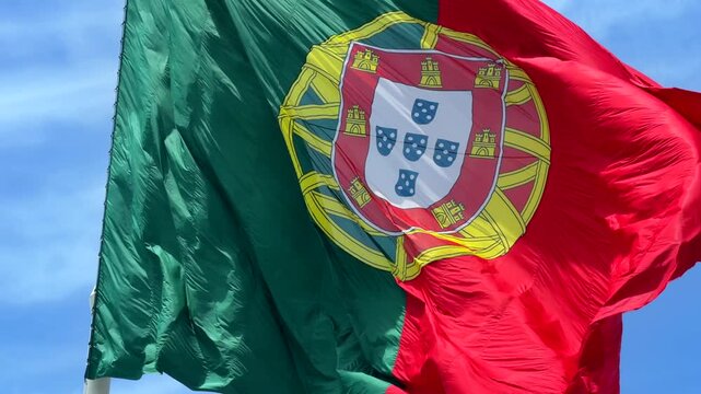Low-angle view of national flag fluttering in strong wind, red and green fabric with central yellow emblem and partial white shield, waving across bright blue sky with soft clouds