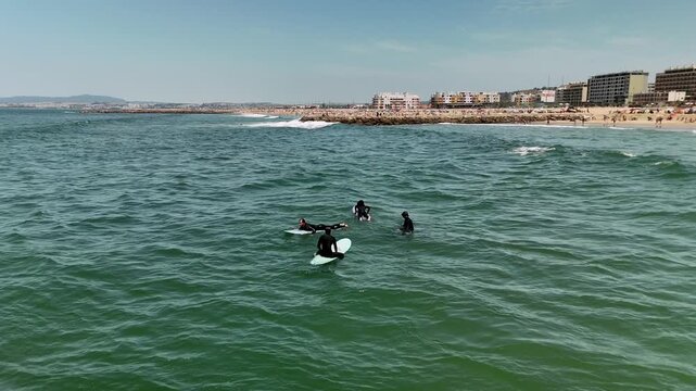 Aerial view of surfers waiting near breaking waves on crowded sandy beach under bright midday sun with umbrellas and low-rise buildings along coastline