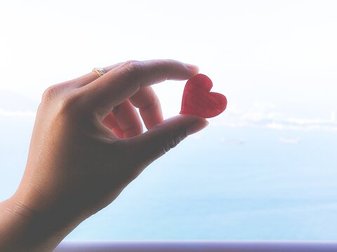 Red heart held aloft in hand against blue sky background. Suitable for love, romance, valentines day, hope, happiness themes.