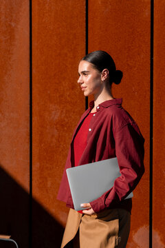 Woman carrying laptop beside rust colored architectural wall