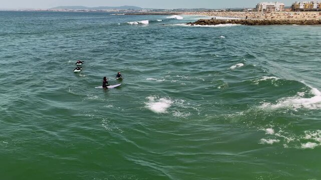 High oblique aerial view of four surfers paddling and waiting on surfboards in emerald green ocean beside sandy coastline and rocky jetty with small breaking waves and foam trails over rippled water