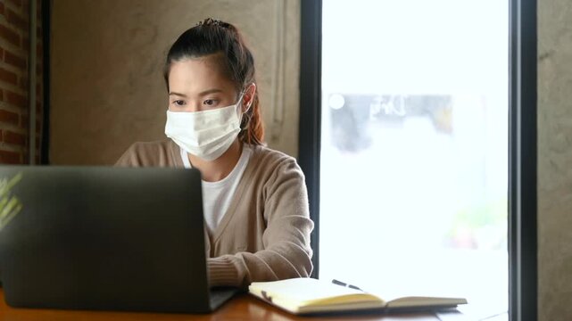 A woman wearing a face mask types on a laptop at a cafe workspace. The woman works beside a notebook and food plate on a wooden table, with window light falling across the brick wall behind her.