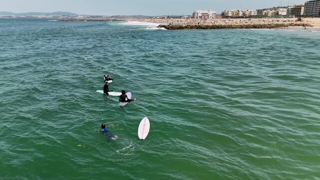 High overhead surf scene with surfers waiting on boards and swimmer near leash lines in green coastal water beside sandy beach and rocky jetty under bright midday sky