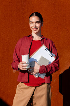 Smiling woman holds coffee and documents by sunlit metal wall