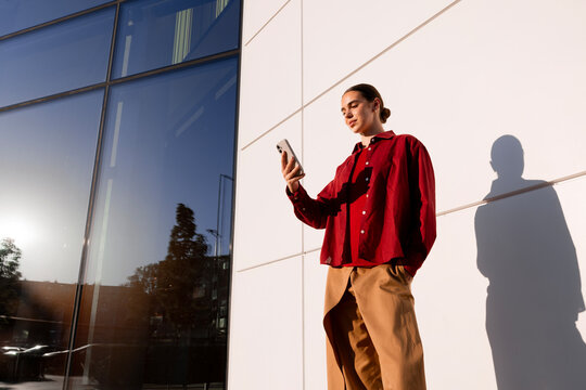Woman checking smartphone beside modern building in afternoon sun