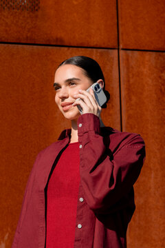 Woman talking on smartphone beside sunlit rust metal wall