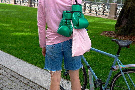 Woman stands near bike with boxing gloves and tote bag in green park