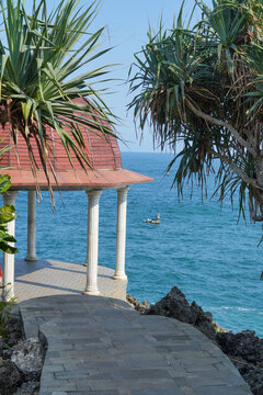 Domed gazebo with classic white pillars on a rocky seaside cliff overlooking the calm blue ocean and clear sky.