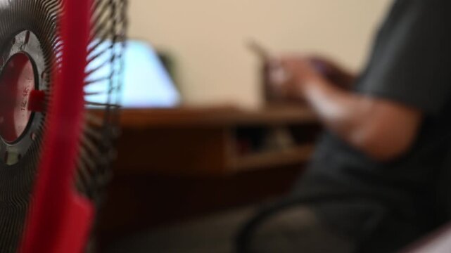 Close-up of an oscillating electric fan rotating in the foreground, with a blurred person working at a computer and using a smartphone in a dim home workspace, illustrating indoor cooling and everyday