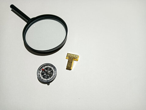 Magnifying glass and compass arranged with a small gold T-shaped object on a white surface, representing exploration, navigation, investigation, discovery, and finding direction.