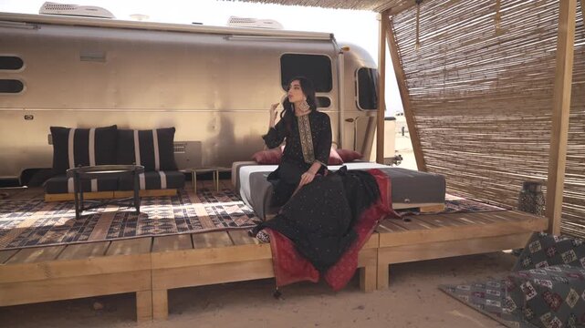 Cinematic shot of an elegant woman relaxing on a wooden deck outside a retro camper trailer in a desert glamping camp