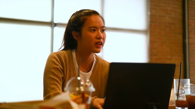 A woman gestures in conversation with another person across a cafe table. The woman faces the other diner as a laptop, cold drinks, glassware, and brick walls surround the wooden table.