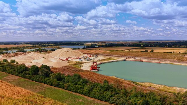 Quarry lake and sand processing plant beneath clouds. A red floating structure connects to shoreline equipment beside green water and banks bordered by vegetation.