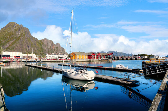 Sailboat at a pier in Svolvaer harbor in the Lofoten Islands with calm water, colorful houses and distinctive mountains beneath a clear blue sky.