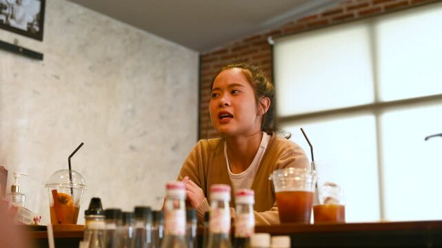 A woman gestures in conversation with another person across a cafe table. The woman faces the other diner as a laptop, cold drinks, glassware, and brick walls surround the wooden table.