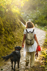 Fototapeta premium A back view of a woman with a backpack walking her black dog along a dirt path through a sunny autumn forest