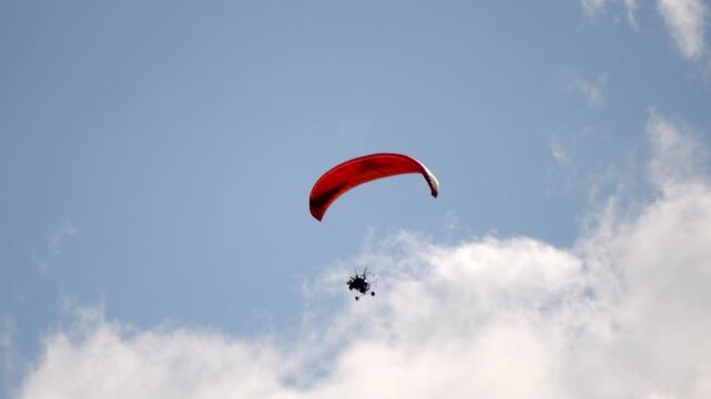 Red parachute glides through blue sky. Person soars above fluffy white clouds. Sky is clear, with no visible obstacles. Joyful thrill seeker floats with open eyes. Ideal for adventure, sports